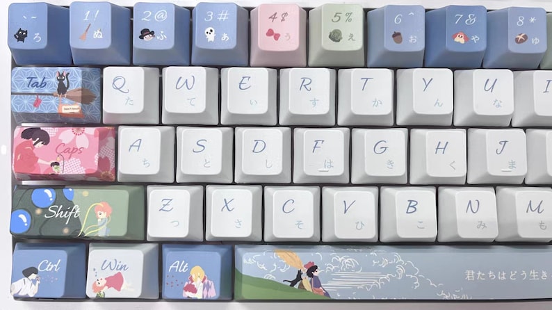 May include: A white mechanical keyboard with blue and pink keycaps featuring Japanese characters and illustrations. The keyboard has a row of function keys with numbers and symbols, a row of letter keys, and a row of modifier keys. The keyboard also has a row of keys with Japanese characters and illustrations. The keyboard has a blue and white space bar with the text "君たちはどう生き" in Japanese.