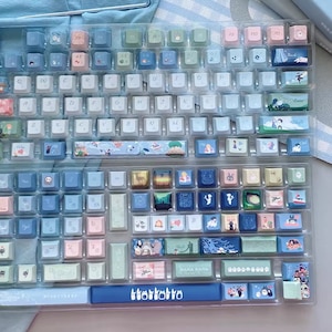 May include: A custom keyboard with a blue and white color scheme. The keys have different designs, including a blue and white striped design, a floral design, and a design with a cat on it. The keyboard also has a blue space bar with the word "TokoToko" printed on it.