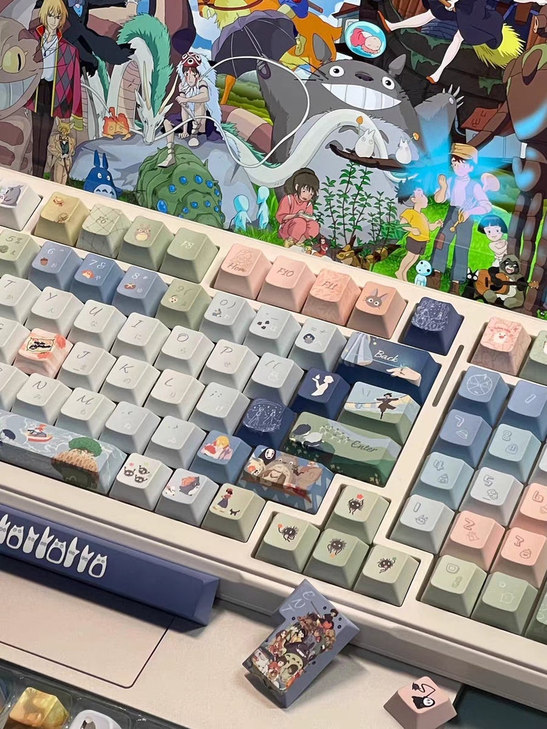 May include: A white mechanical keyboard with pastel blue and pink keycaps featuring characters from the animated film 'My Neighbor Totoro'. The keyboard has a blue and white space bar with the word 'ortotro' printed on it.
