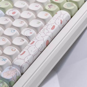 Puede incluir: Primer plano de un teclado mecánico blanco con teclas personalizadas. La barra espaciadora presenta un patrón repetido de personajes de dibujos animados con acentos rosas. Otras teclas son blancas con símbolos y letras en colores pastel.