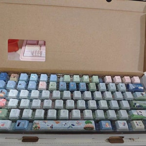 May include: A white keyboard with a variety of pastel colored keycaps. The keycaps have different designs, including flowers, animals, and landscapes. The keyboard is in a cardboard box.