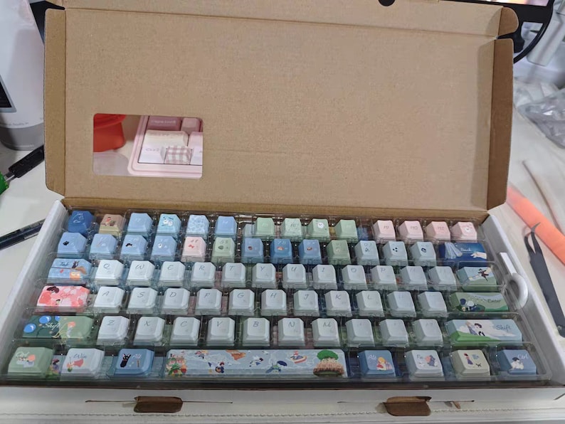 May include: A white keyboard with a variety of pastel colored keycaps. The keycaps have different designs, including flowers, animals, and landscapes. The keyboard is in a cardboard box.