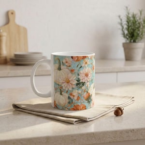 Könnte beinhalten: Ein weißer Keramikbecher mit einem wiederkehrenden Muster aus hellen Kürbissen, zartrosa Dahlien und orangen Blumenakzenten auf hellblauem Grund. Der Becher hat einen geschwungenen Henkel und steht auf einer gefalteten beigen Stoffserviette.