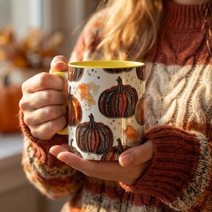 Könnte beinhalten: Ein Keramikbecher mit weißem Hintergrund und einem wiederkehrenden Muster aus orangefarbenen und braunen Kürbissen mit detaillierten Linien und orangefarbenen Aquarellspritzern. Der Becher hat ein leuchtend gelbes Inneres und einen passenden gelben Henkel.