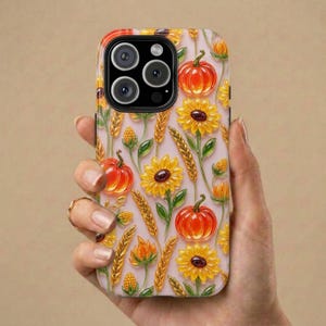Könnte beinhalten: Eine Handyhülle mit einem wiederkehrenden Muster aus orangefarbenen Kürbissen, gelben Sonnenblumen und goldenen Weizenähren auf hellrosa Hintergrund. Das Design umfasst grüne Stängel und Blätter sowie einen schwarzen Kameraausschnitt.