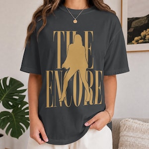 Puede incluir: Una camiseta de manga corta gris carbón con un estampado gráfico dorado. El diseño muestra el texto THE ENCORE en una fuente serif grande, con la silueta de una persona con chaqueta y tacones altos superpuesta sobre las letras.