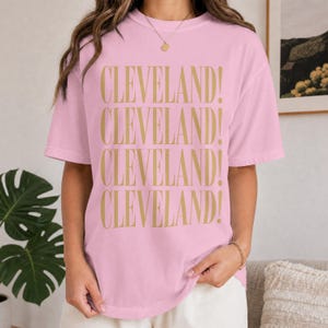 Könnte beinhalten: Ein hellrosa T-Shirt mit kurzen Ärmeln, auf dem das Wort CLEVELAND! viermal in einer vertikalen Spalte in einer goldenen Serifenschrift gedruckt ist. Das Kleidungsstück hat einen Rundhalsausschnitt und eine lockere Passform.