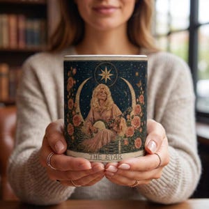 Op de afbeelding: Een keramische mok met een illustratie in tarotstijl van een blond persoon die banjo speelt, omringd door roze rozen en een halve maan tegen een donkerblauwe sterrenhemel, met de tekst THE STAR onderaan het ontwerp gedrukt.