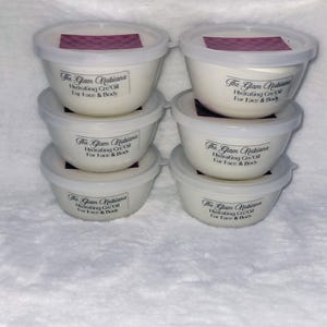 Puede incluir: Seis recipientes blancos con tapas, cada uno etiquetado "The Glam Nubiana Hydrating Cre-Oil For Face & Body". El fondo es una superficie rosa acolchada. Para rostro y cuerpo.