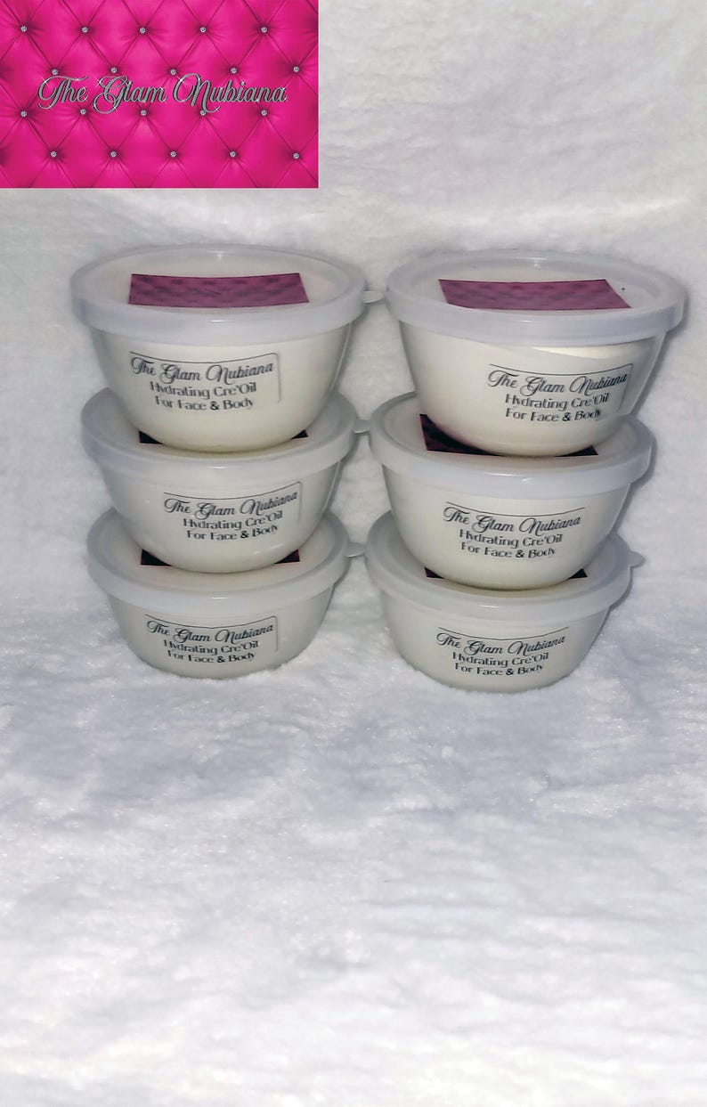Puede incluir: Seis recipientes blancos con tapas, cada uno etiquetado "The Glam Nubiana Hydrating Cre-Oil For Face & Body". El fondo es una superficie rosa acolchada. Para rostro y cuerpo.
