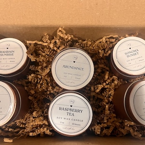 Könnte beinhalten: Ein Set mit sechs Sojawachskerzen in braunen Metalldosen mit weißen Etiketten. Die Kerzen sind in einer Box mit braunem Wellpapier angeordnet. Die Etiketten zeigen die Namen der Düfte: Bahrain Sunrise, Abundance, Sonoma Sunset, Aurora, Raspberry Tea und Ruby Kisses.