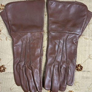 May include: A pair of brown leather gloves. The gloves have a classic design with a long cuff and detailed stitching on the fingers. The leather has a smooth, polished finish.