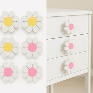 May include: White daisy-shaped drawer pulls with yellow or pink centres. The pulls are on a white background and attached to a white chest of drawers with three drawers. The daisy pulls add a whimsical touch to furniture.