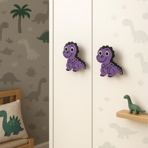 May include: Two purple dinosaur cabinet knobs with black trim are fixed to a white cabinet. The knobs have large, friendly eyes. The background features dinosaur-themed wallpaper and a small green dinosaur figurine on a shelf. A dinosaur pillow is on a wooden chair.