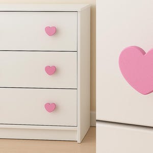 May include: A white chest of drawers with three drawers, each featuring a pink heart-shaped knob. A close-up shows a single pink heart-shaped knob on a white surface. The knobs add a playful touch to the furniture.