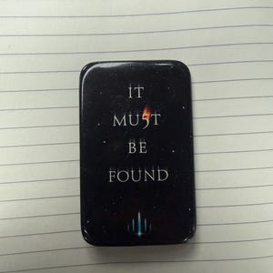 Peut inclure: Une épingle noire rectangulaire aux coins arrondis. L'épingle présente le texte blanc « IT MUST BE FOUND » avec une petite flamme et un graphique de fusée. Le fond est un ciel étoilé.