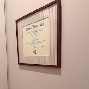 Könnte beinhalten: Ein Master of Public Health Diplom der Drexel University für Lindsay Dunbar, präsentiert in einem rechteckigen Holzrahmen mit weiß-grau gestreiftem Passepartout. Das Dokument enthält schwarzen Text, offizielle Unterschriften und ein blaues Siegel.
