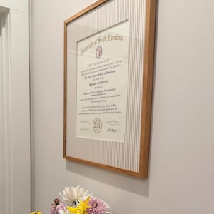 Może przedstawiać: Dyplom University of South Carolina w jasnej drewnianej ramie z białym passe-partout w pionowe prążki. Dokument zawiera nazwę uczelni, szczegóły stopnia naukowego oraz podpisy, umieszczony na gładkiej ścianie.