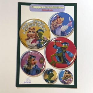 Op de afbeelding: Een knoppenset met Muppets-personages in afstudeerkleding. De ronde knoppen tonen Miss Piggy en Kermit de Kikker. Tekst op de knoppen is onder meer "Have you hugged a graduate today?" en "Brains are beautiful!"