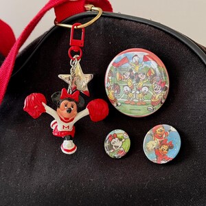 May include: A black canvas bag adorned with a Minnie Mouse cheerleader keychain featuring red pom-poms and a white uniform with an M, a large circular pin showing Disney characters cheering, and two smaller circular pins depicting Minnie Mouse in red.