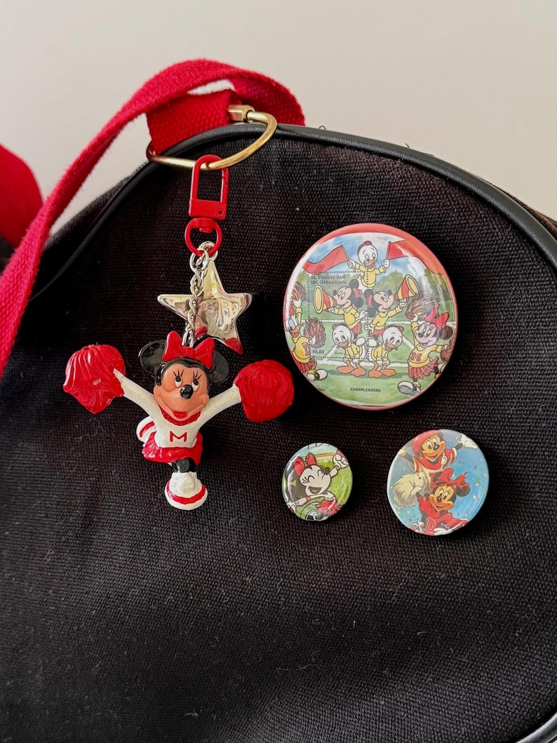 May include: A black canvas bag adorned with a Minnie Mouse cheerleader keychain featuring red pom-poms and a white uniform with an M, a large circular pin showing Disney characters cheering, and two smaller circular pins depicting Minnie Mouse in red.