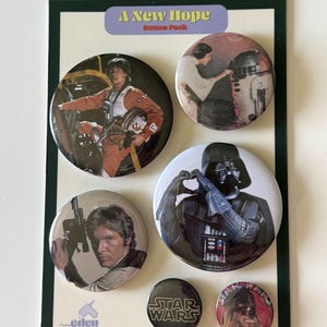 May include: A collection of six Star Wars themed button pins. The pins feature characters and logos from the movie, including Luke Skywalker, Princess Leia, R2-D2, Darth Vader, Han Solo, Chewbacca, and the Star Wars logo. The pins are mounted on a green and black backing.