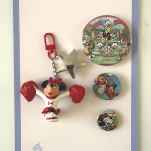 May include: A keychain featuring a Minnie Mouse cheerleader figure with red pom-poms and a silver star charm, accompanied by three circular pins. The largest pin depicts Disney characters cheering, while the two smaller pins show Minnie Mouse.