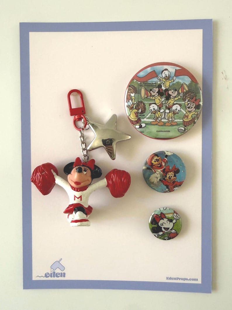 May include: A keychain featuring a Minnie Mouse cheerleader figure with red pom-poms and a silver star charm, accompanied by three circular pins. The largest pin depicts Disney characters cheering, while the two smaller pins show Minnie Mouse.
