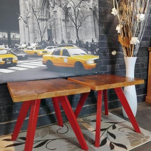 May include: Two square side tables with thick, natural-toned wooden tops and four angled, bright red metal legs each. The tables feature a simple, sturdy construction with a smooth finish on the wood surfaces and a solid colour on the metal legs.