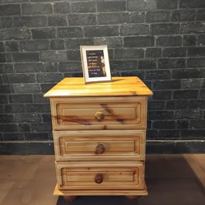 May include: A three-drawer wooden bedside table with a light brown finish. The table has rounded wooden knobs and sits on small, rounded feet. A framed picture rests on top of the table.