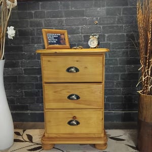 May include: A three-drawer wooden bedside table in a light brown finish. It features black metal handles and a decorative base. A small framed picture and an alarm clock are on the top surface.