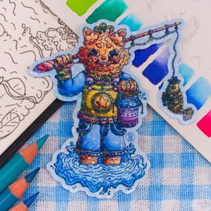 Op de afbeelding: Een glittersticker met een oranje, katachtig wezen dat in blauw water staat. Het wezen draagt een geel vest met een ster, een blauwe broek en houdt een hengel met een kleine vis aan de lijn en een paarse emmer in de andere hand vast.