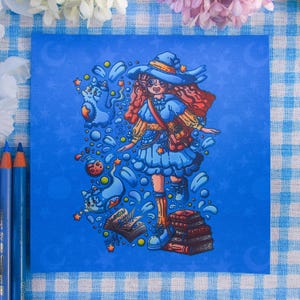 Puede incluir: Una impresión artística cuadrada sobre fondo azul que muestra a un personaje con sombrero de bruja azul, vestido azul con mangas amarillas y un bolso. El personaje está rodeado de espíritus felinos azules flotantes, una poción y un libro abierto.