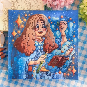 May include: A square art print featuring a cartoon character with long wavy brown hair, wearing round glasses and a blue outfit. The character holds a green potion bottle and a small strawberry, with a floating open book surrounded by blue swirls.