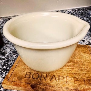 May include: Cream-colored mixing bowl with a pouring spout, resting on a wooden cutting board. The cutting board has the words "BON APPETIT" engraved on it. The bowl is a kitchenware item.
