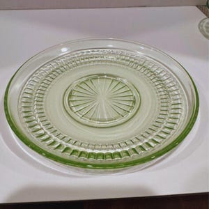 May include: A clear, light green glass plate with a circular design. The plate features a raised, ridged border and a central, sunburst-like pattern. The plate is likely vintage and suitable for serving food.