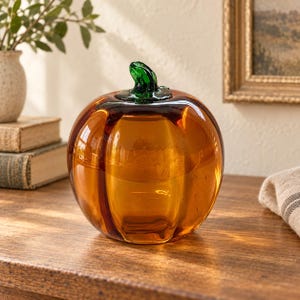 May include: A decorative glass pumpkin sculpture featuring a smooth, translucent amber-colored body with vertical ridges and a distinct, glossy dark green glass stem attached to the top, creating a reflective surface with visible light highlights.