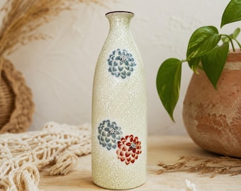 Puede incluir: Un jarrón alto de cerámica blanquecina con cuello estrecho y superficie texturizada. El jarrón está decorado con dos diseños florales azules y uno rojo. El jarrón mide aproximadamente 25 cm de altura.