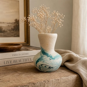 May include: A ceramic vase with a textured cream-colored upper section and a smooth, rounded base featuring a marbled pattern of teal, blue, and white swirls. The vase contains a cluster of delicate, dried white baby's breath flowers.