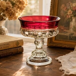 Puede incluir: Un cuenco de cristal sobre pedestal con un borde rojo intenso y una base transparente con un patrón de relieve texturizado. El cuenco tiene una abertura circular ancha y un tallo robusto de varios niveles sobre una base redonda.