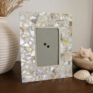 May include: A rectangular photo frame with a mosaic design made of iridescent, irregularly shaped pieces. The frame's interior has a grey backing with two black dots. The frame is on a dark brown surface, with a vase and seashells in the background.
