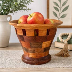 Op de afbeelding: Een houten fruitschaal met een vlechtwerkpatroon, gevuld met rode en gele appels. De schaal is gemaakt van verschillende bruintinten en staat op een tafel. Op de achtergrond staat een vaas met groen.