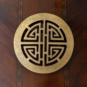 May include: A circular brass trivet featuring an openwork geometric pattern in the centre, surrounded by a decorative border of engraved flowers and leaves. The metal has a warm, golden tone with visible signs of age and patina throughout the surface.