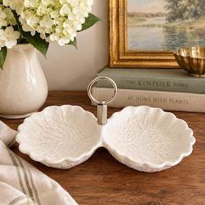 May include: A white ceramic double-bowl serving dish with a textured leaf-vein pattern on the interior and exterior. The two bowls are connected by a central silver-toned metal post featuring a circular ring handle at the very top.