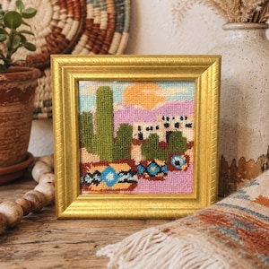 May include: A framed needlepoint artwork featuring a desert landscape with green cacti, a setting sun, and a pink and blue sky. The artwork is in a gold-colored frame and is displayed on a wooden surface.
