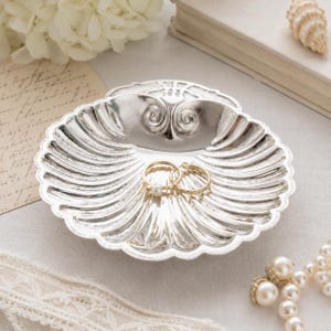 May include: A silver-toned, shell-shaped trinket dish with a scalloped edge and a decorative spiral detail at the top. Two gold-toned rings, one featuring a small white pearl, rest inside the center of the dish, which has a polished finish.