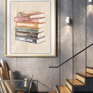 May include: A framed print featuring a stack of books with titles like "I Smell Snow" and "Meet Me in Stars Hollow." The books are in shades of brown, pink, and orange, set against a beige background. The frame is a light wood color, and the print is displayed on a gray wall.