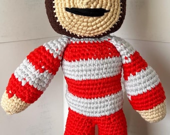 May include: A crocheted plush toy in the shape of a person wearing a red and white striped shirt, red pants, and a white hat with a red band. The toy has brown hair, a beige face, and black eyes.