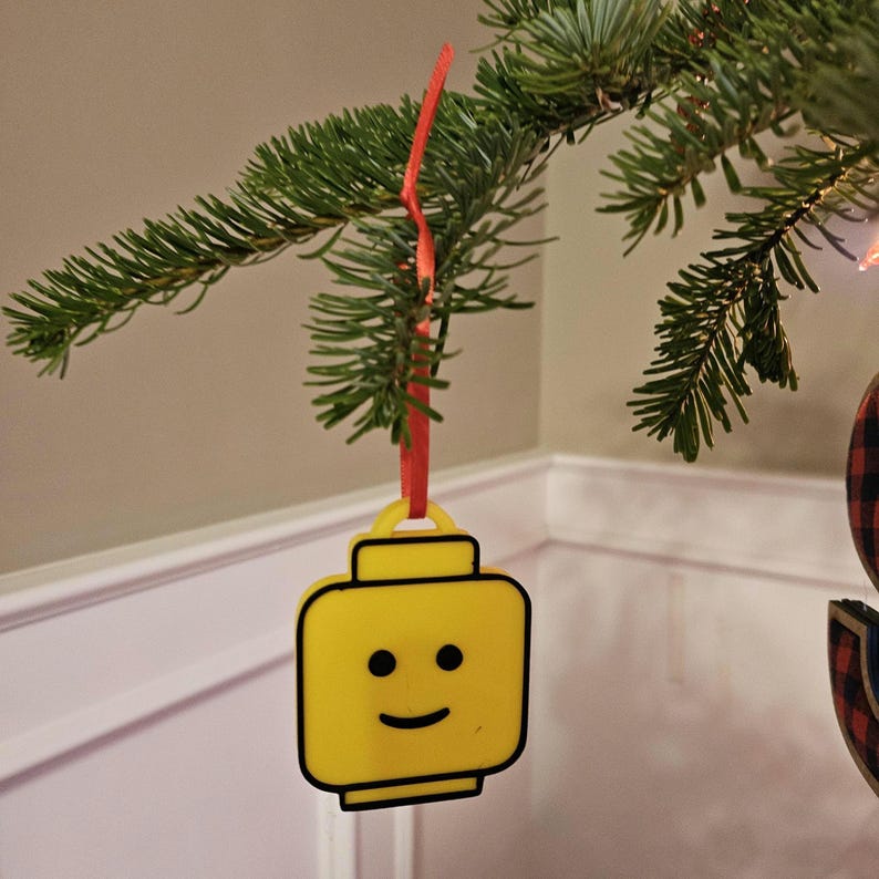 May include: A yellow Lego minifigure head ornament with a smiling face, hanging from a red ribbon on a Christmas tree.