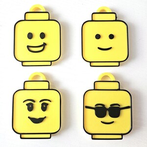 May include: Four yellow plastic Lego minifigure keychains with different facial expressions. The minifigures have black outlines and black facial features. One minifigure has a smile, one has sunglasses, one has a surprised expression, and one has a neutral expression.